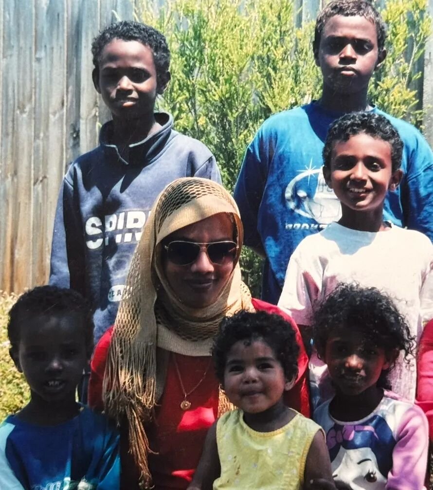 A woman wearing sunglasses and head scarf seated with her six young children around her.