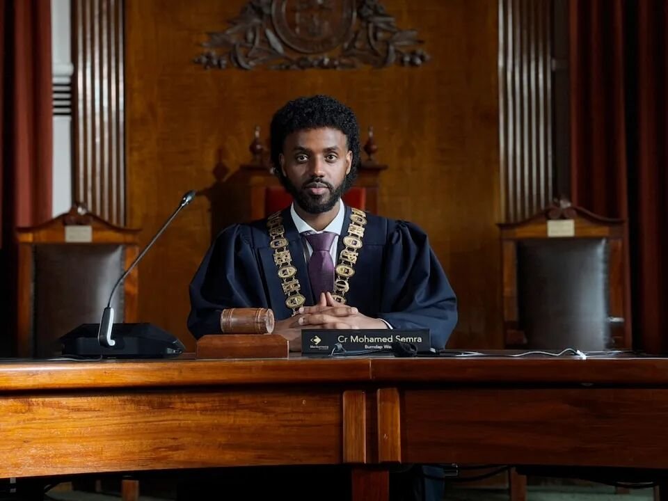Mohamed Semra wears black mayoral robes and chain while seated at solid timber chambers desk, his hands clasped.