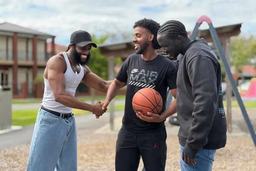 Three young men stand together smiling on a basketball court. One is holding a basketball and shaking hands with another.