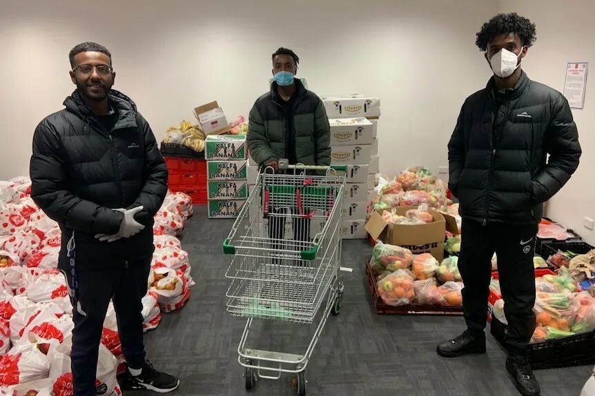 Three young men stand a distance apart in a room that has grocery bags and trolley in centre. All wear face masks.