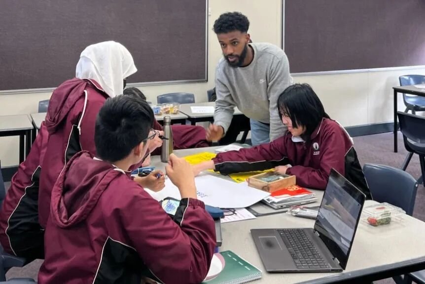 A man with facial hair leans over a cluster of school desks to chat with three high school students in uniform.