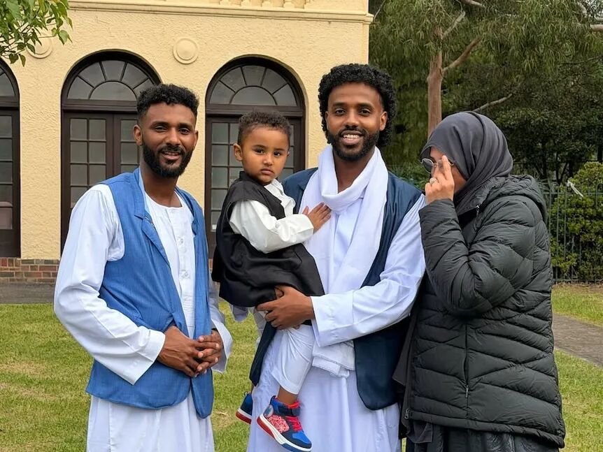 Two men and a woman stand in front of a building outdoors. One man (centre) is holding a toddler.