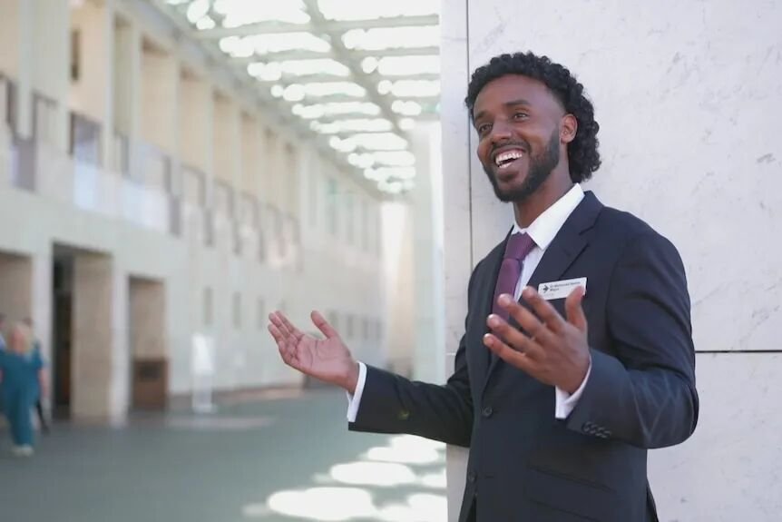 Man wearing a suit stands smiling with his hands out. A corporate building is in the background.